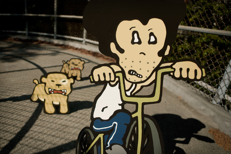 two cardboard dogs chasing a cardboard man on a bicycle